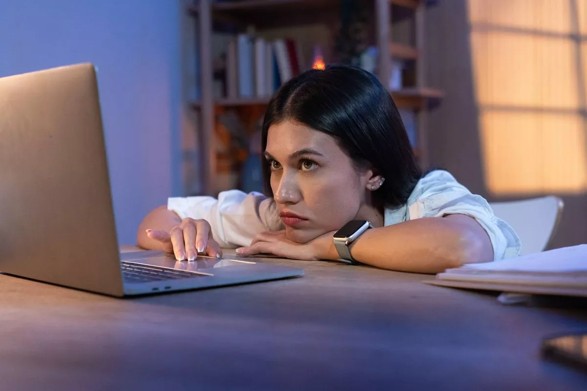 A young professional woman appears exhausted and contemplative as she works on her laptop late into the night, signaling dedication and overtime work.