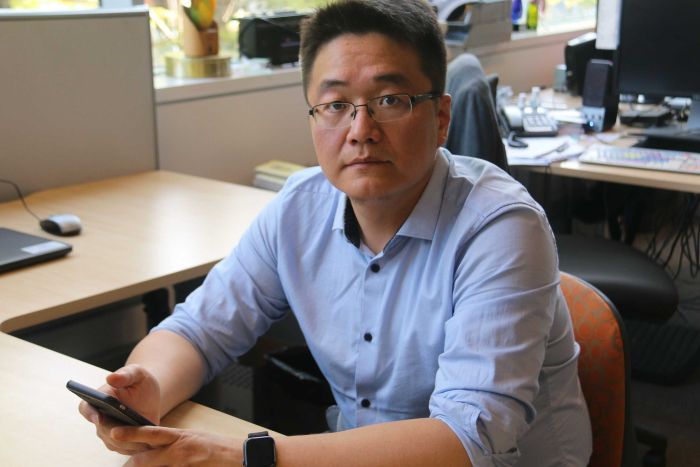 James Jing A man sits at a desk holding a mobile phone.