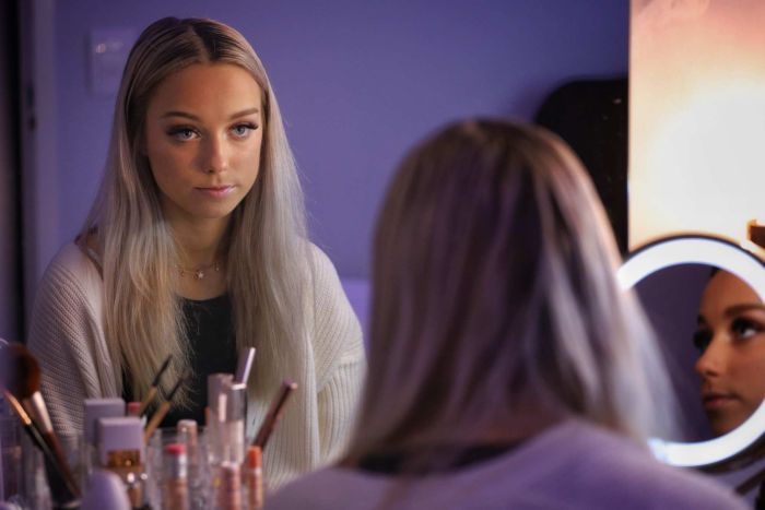 TikToker Olivia Plant Olivia Plant looking into a mirror with long blonde hair and wearing a white cardigan, sitting at a table with make-up items