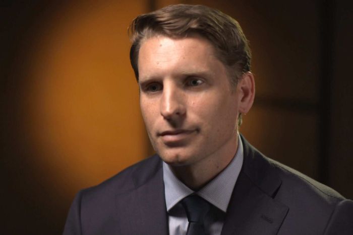 Andrew Hastie Head shot of Andrew Hastie wearing grey suit and dark tie, with circle of orange light on the wall behind him