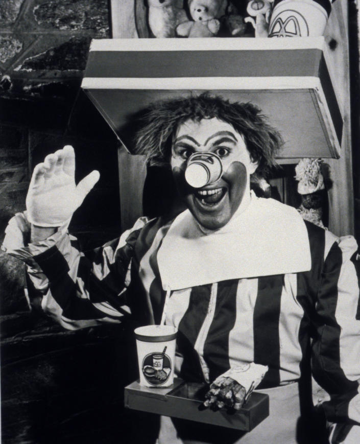 Man in a clown costume waving and smiling with a McDonald's cup and fries in front of him