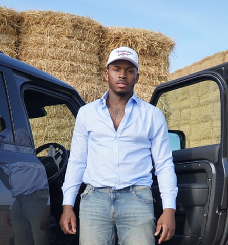 Fekky wearing a shirt and white baseball cap standing in front of a large haystack.