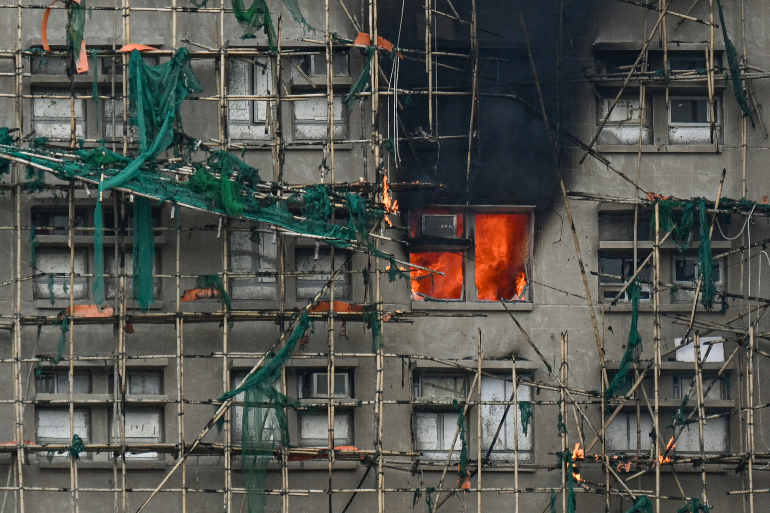 Apartment windows on fire within a building surrounded by scaffolding and green netting in Hong Kong.
