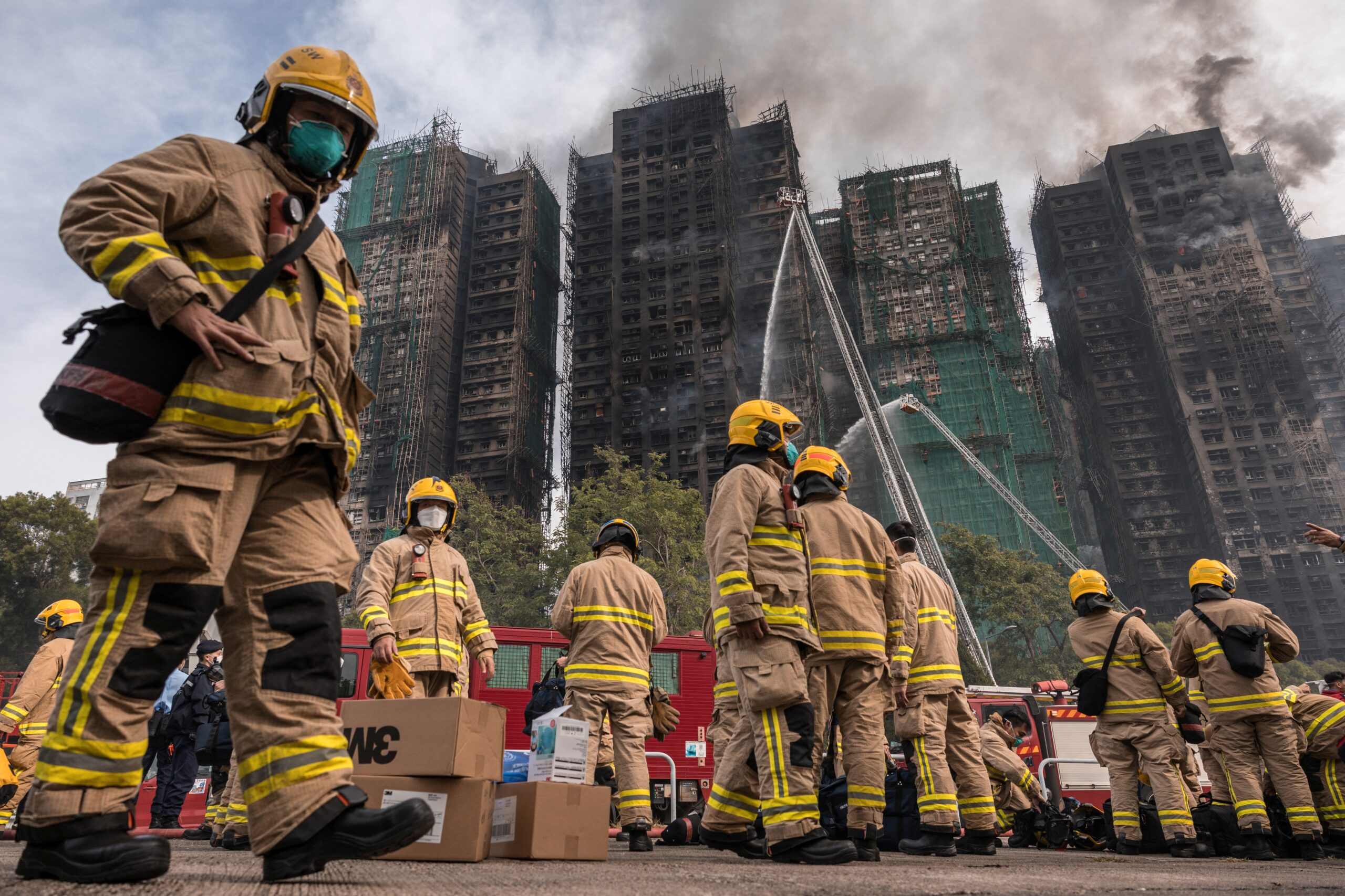 Firefighters in tan suits and yellow helmets prepare at the scene of a fire that destroyed multiple apartment buildings.