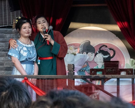 Granny Wang on stage with her arm around a young woman who wears a pale blue flower-patterned dress; they stand in front of a large cartoon showing an anime-style cute, happy couple