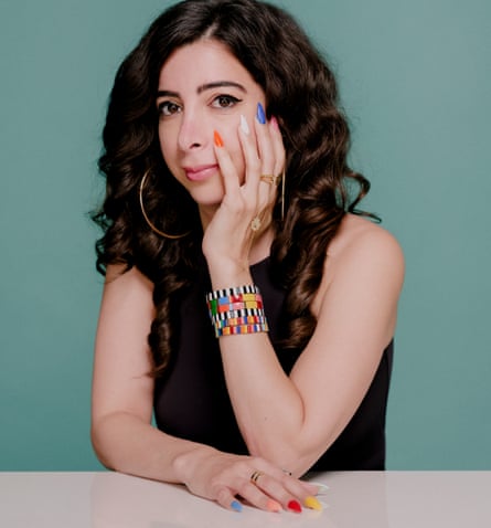  Zahra Hankir, sitting at a table, with one hand cupping her her face and the other on the table, each long nail painted a different bright colour