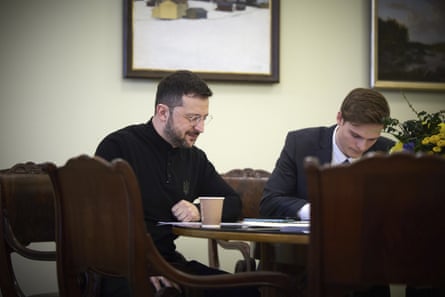 A smiling man sits at a table with a cup and papers on it