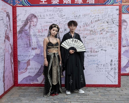 Zhang Hongqian and Hongguang stand in front of a wall with scribbled names and numbers; she wears a cropped top and long skirt with elaborate embroidery plus a headdress, and he wears a long black kimono-style gown and carries a white fan