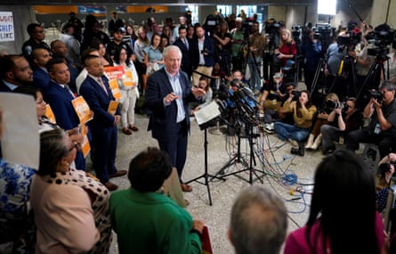 man wearing suit stands behind microphone as crowd of people listen to him speak