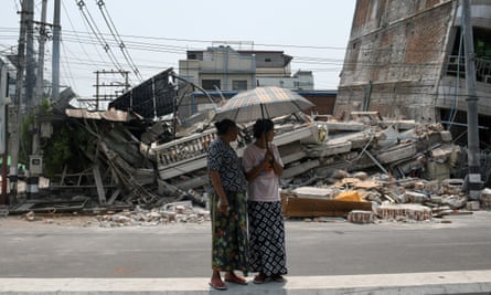 Two women stand under a parasol in front of collapsed buildings