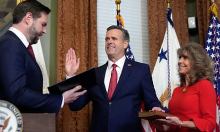 A man holds his right hand up as his left hand is on the Bible held by a woman; they both face another man reading from a book
