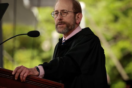 person wearing glasses and black robe stands behind podium and small microphone