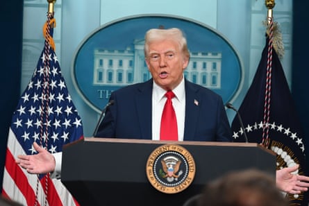 man wearing navy suit and red tie gestures while standing behind podium