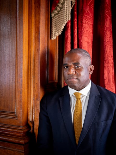 Portrait of David Lammy in a dark suit and yellow tie stood in front of heavy red curtains and a wood-panelled wall