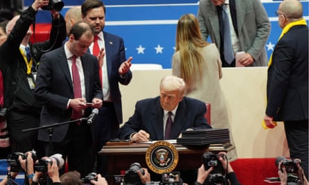 A man signs papers at a desk as people gather around him and a crowd takes photos