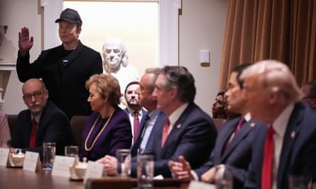 a man speaks while standing in a room full of people seated at a table