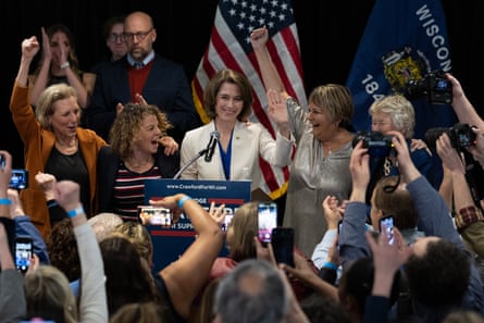 A woman smiles and holds her right hand up at a lectern as people around her cheer and pump their fists in the air