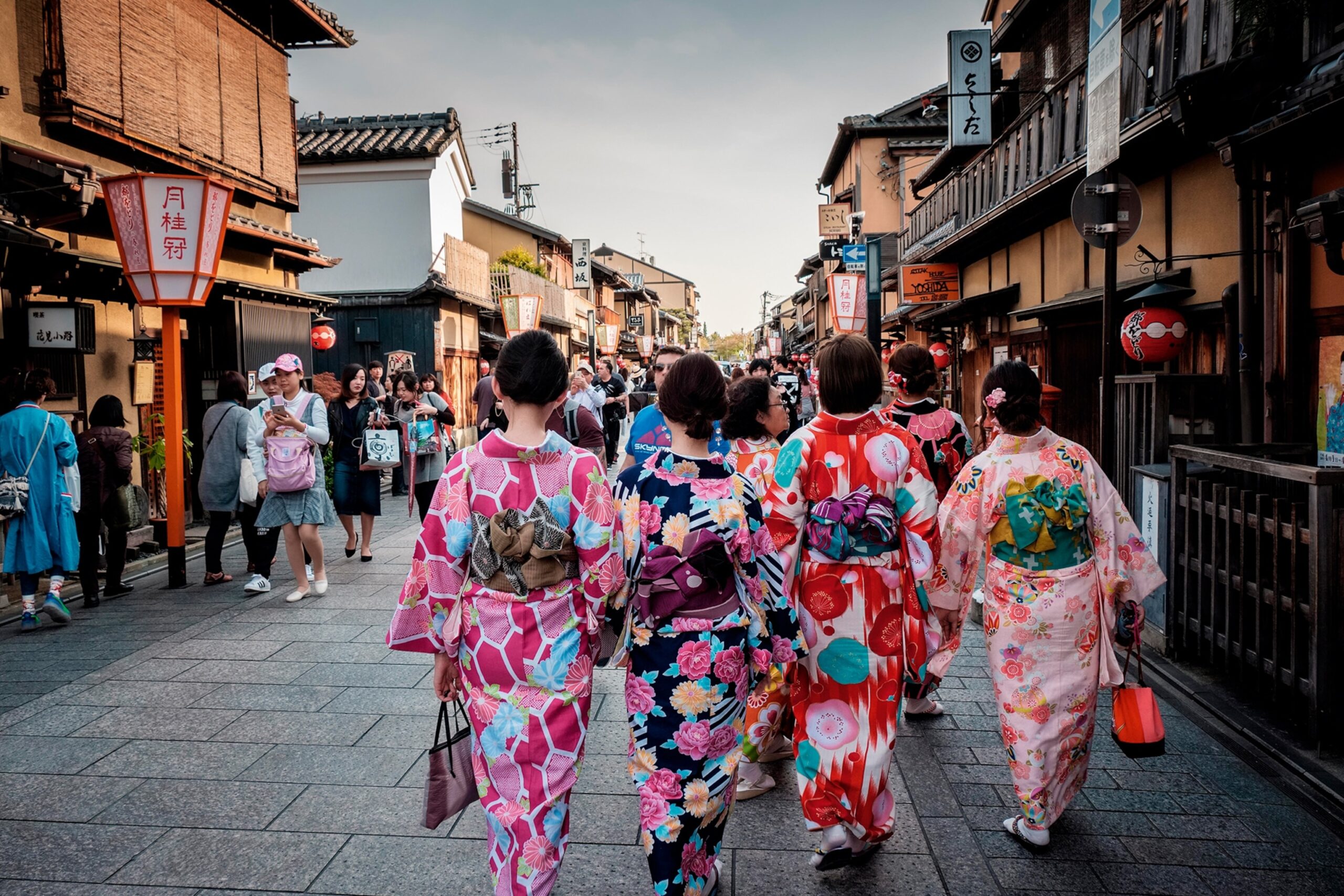 Four traditionally-dressed geishas, seen from behind, walking down a busy street in the ancient town of Kyoto.