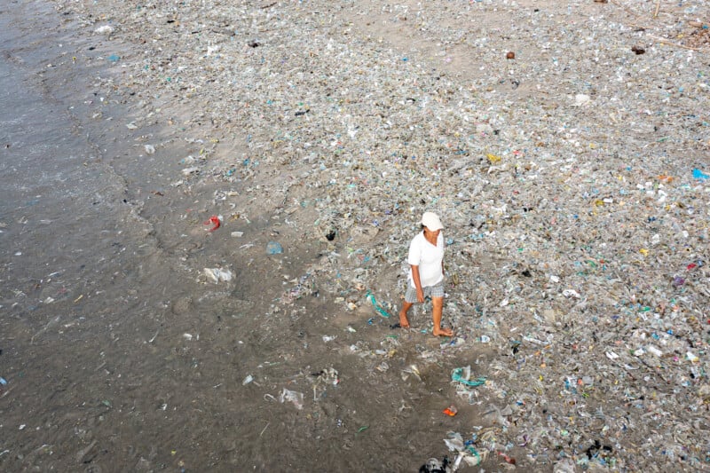 A person wearing a white shirt, shorts, and hat walks barefoot along a beach covered in trash and plastic debris, highlighting severe pollution.
