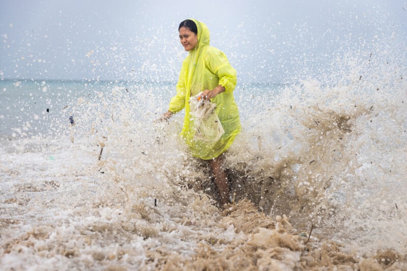A woman in a bright yellow raincoat stands in the ocean as waves splash around her, holding a net and looking determined. The sky is overcast and the water is turbulent.