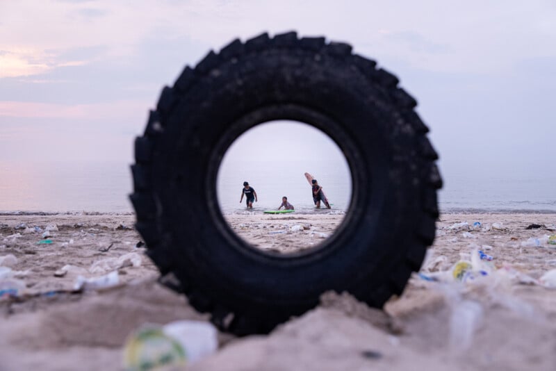 A large tire stands upright on a littered beach, framing three people near the shore through its center; one is holding a surfboard, with the sea and cloudy sky in the background.