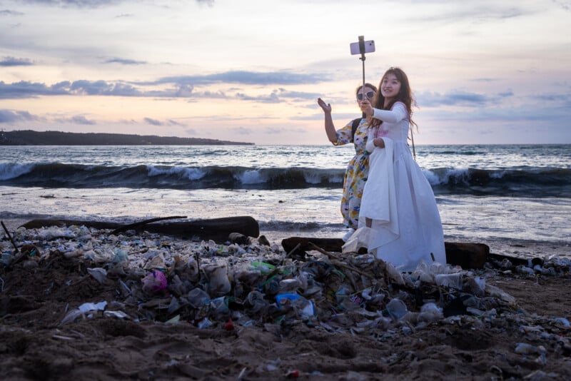 Two women, one in a white dress, take a selfie on a beach littered with trash at sunset. Waves and a cloudy sky are in the background.