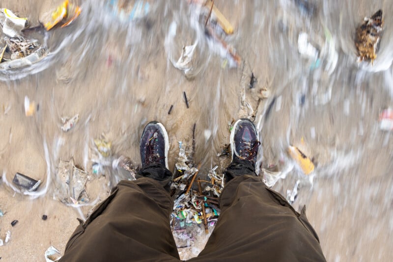 A person stands on a sandy beach, wearing dark pants and sneakers, as waves with floating trash and debris wash over their feet.