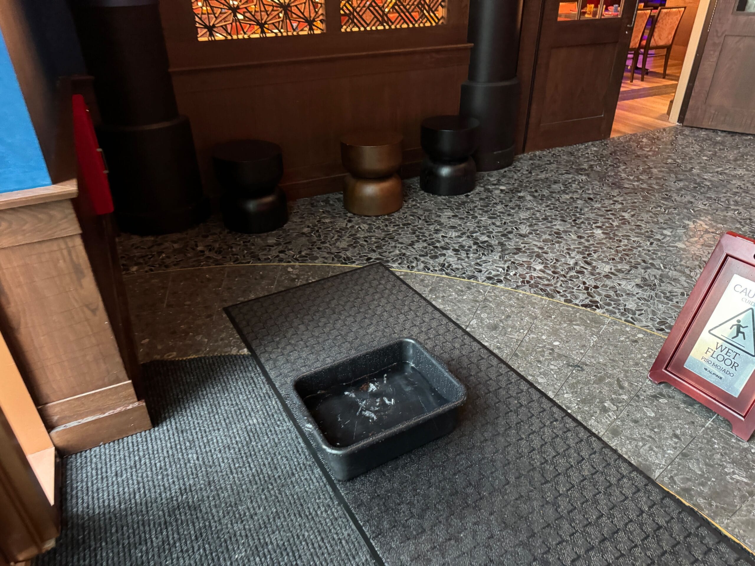 A black plastic bin sits on a mat near a restaurant entrance, with chairs and a wet floor caution sign nearby.