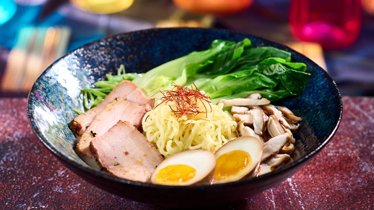 A bowl of ramen with sliced pork, bok choy, mushrooms, marinated eggs, and noodles, garnished with thin red pepper strands—truly an epic universe of flavors in every bite.