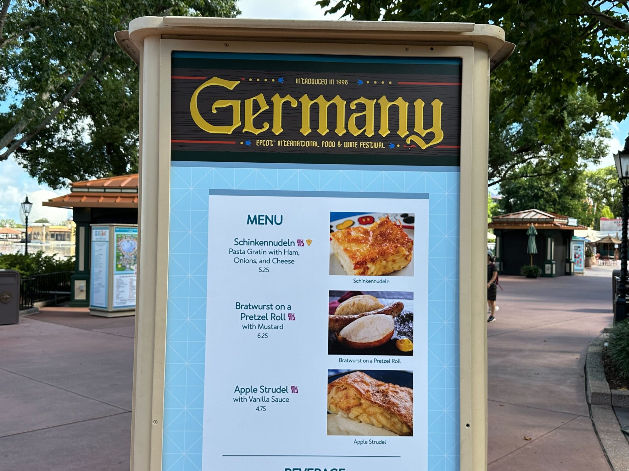 Menus (With Prices) for 2024 EPCOT International Food & Wine Festival 32 A signboard at Epcot's International Food & Wine Festival showcasing the menu for Germany, featuring Schinkenudeln pasta, bratwurst on a pretzel roll, and apple strudel with vanilla sauce.