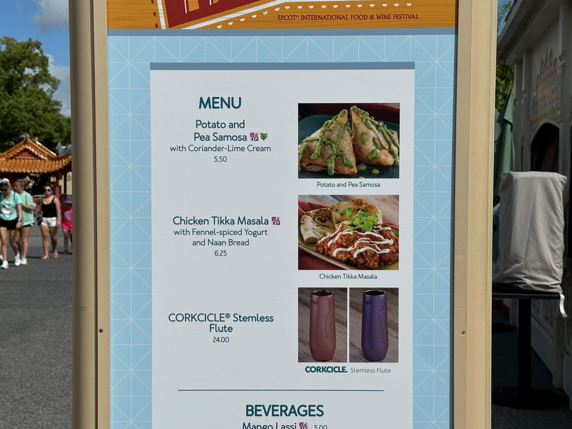 Menus (With Prices) for 2024 EPCOT International Food & Wine Festival 23 A menu sign at the food & wine festival listing items like Potato and Pea Samosa with Coriander-Lime Cream, Chicken Tikka Masala with Fennel-Spiced Yogurt and Naan Bread, Corkcicle Stemless Flute, and a variety of beverages.