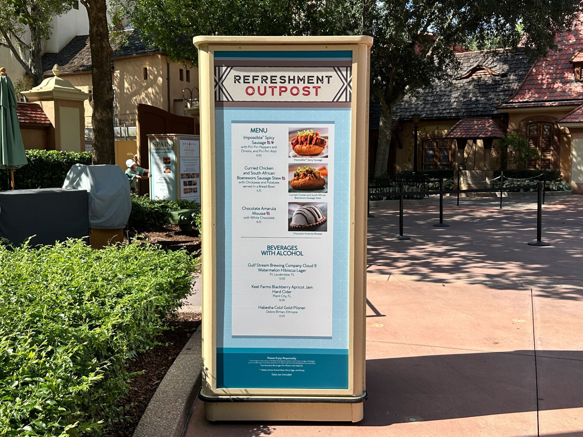 Menus (With Prices) for 2024 EPCOT International Food & Wine Festival 25 Sign at the “Refreshment Outpost” displaying a menu with food items like caramel-coated peanuts, chocolate banana, and beverages, including alcoholic options. Trees and buildings are in the background.