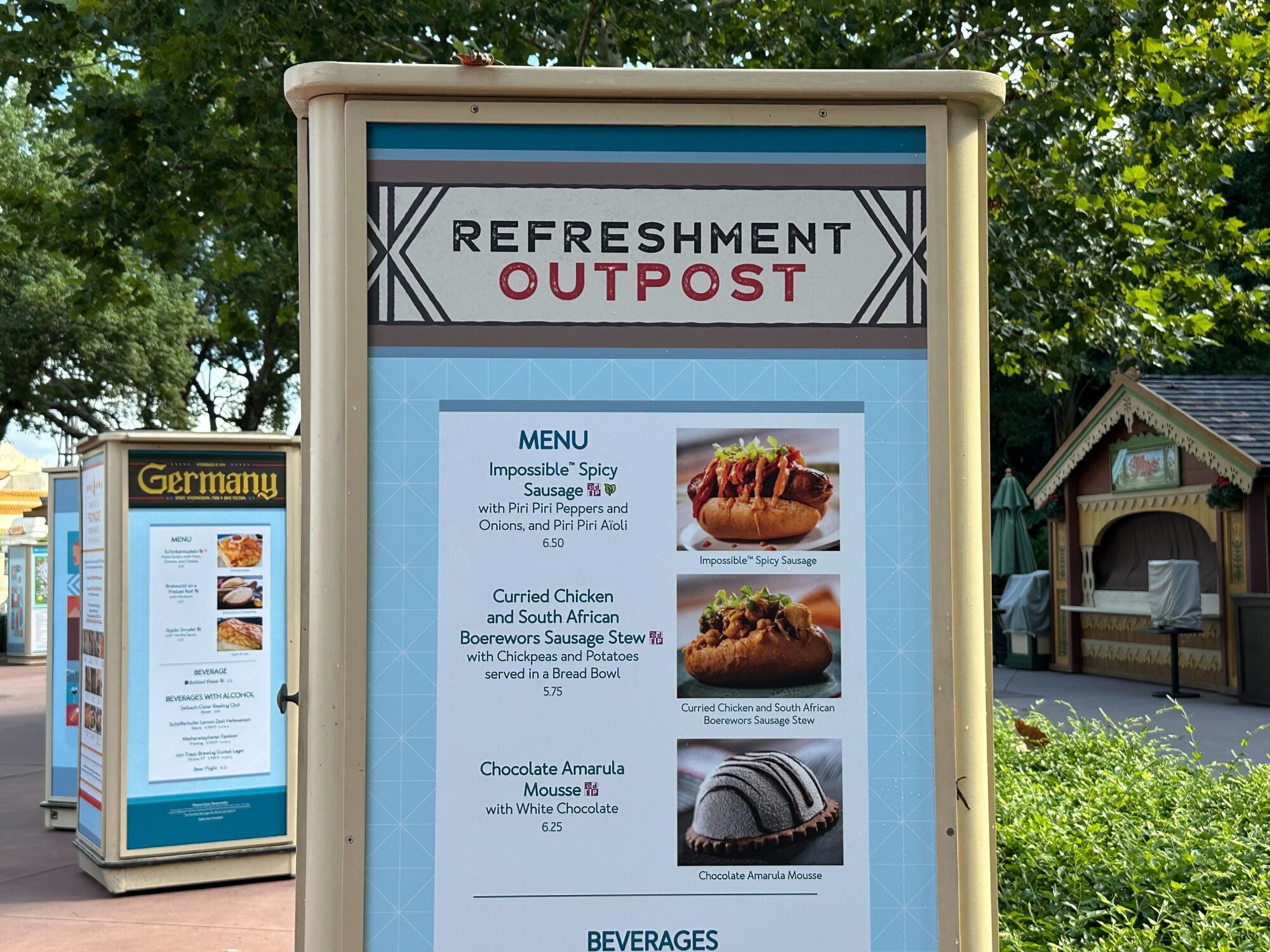 Menus (With Prices) for 2024 EPCOT International Food & Wine Festival 26 A menu at the Refreshment Outpost displaying dishes including Impossible Spicy Sausage, Curried Chicken and South African Boerewors Sausage Stew, and Chocolate Amarula Mousse. Germany sign in background.