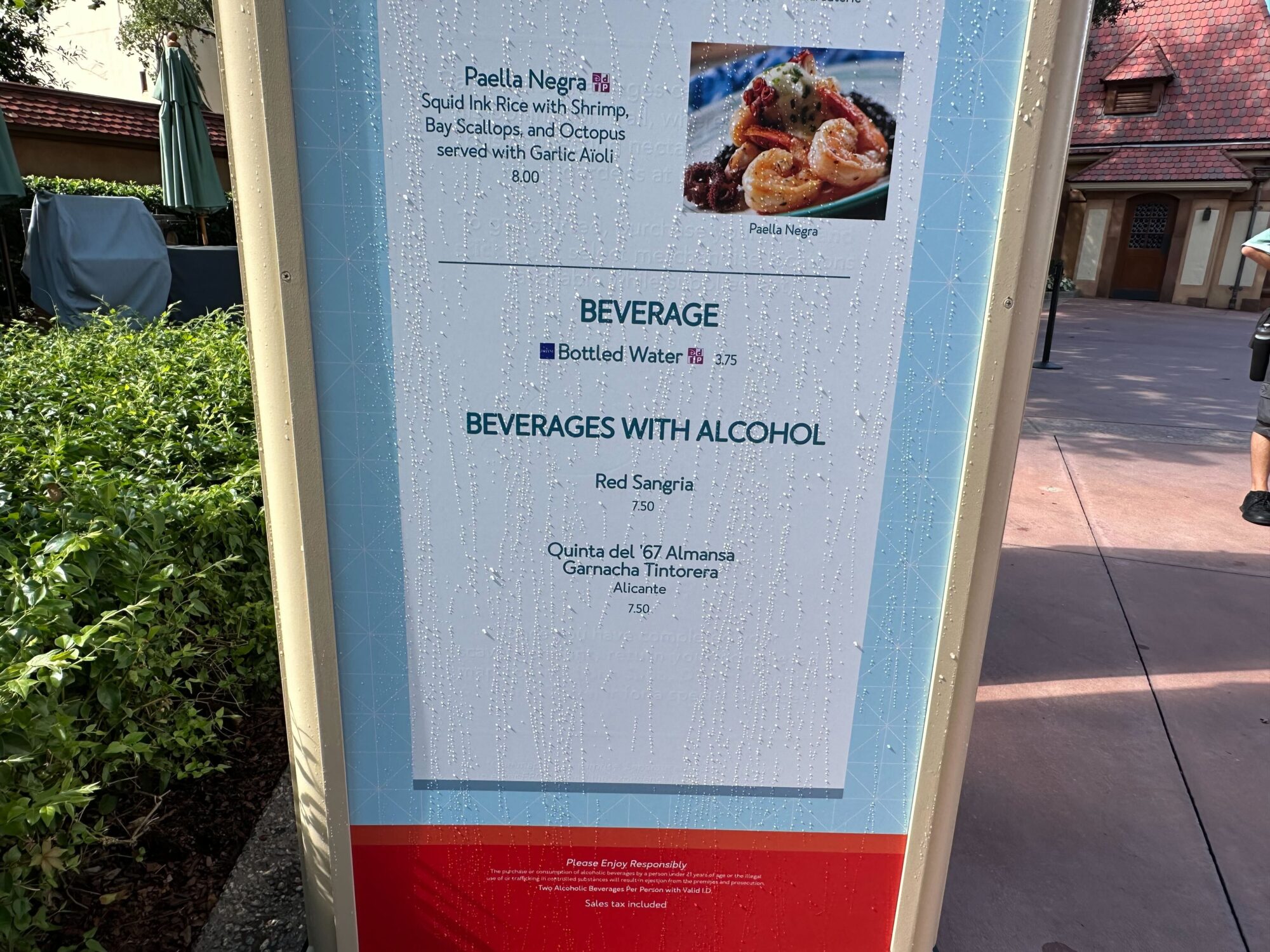 Menus (With Prices) for 2024 EPCOT International Food & Wine Festival 36 Close-up of a menu board displaying food and drink options including Paella Negra, bottled water, red sangria, and wine. The board is slightly wet, likely from rain or condensation.