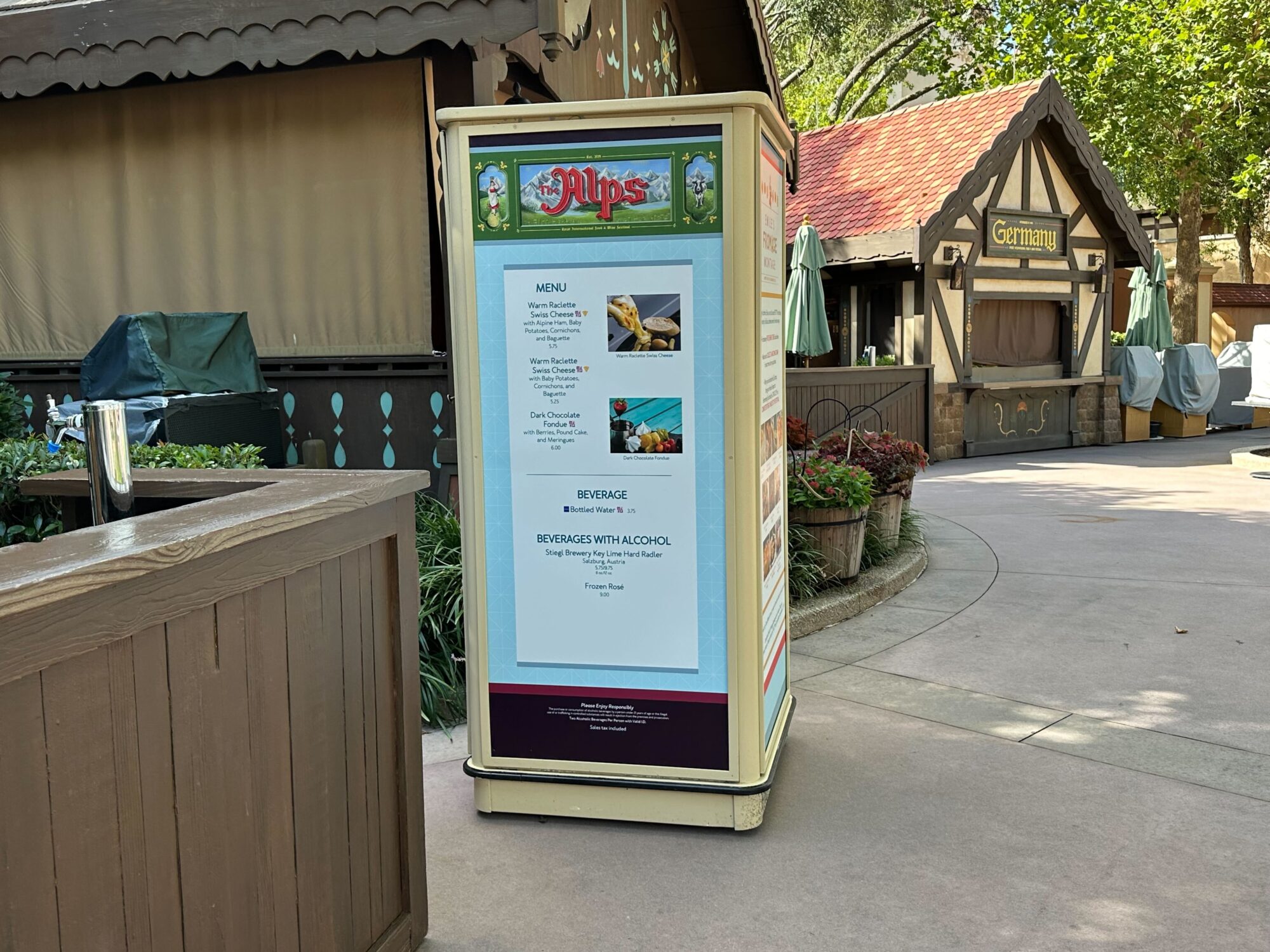 Menus (With Prices) for 2024 EPCOT International Food & Wine Festival 28 A menu board stands outside a rustic food kiosk in an outdoor setting, listing various food and beverage options. Nearby, there's a closed wooden counter and covered items. Trees are in the background.