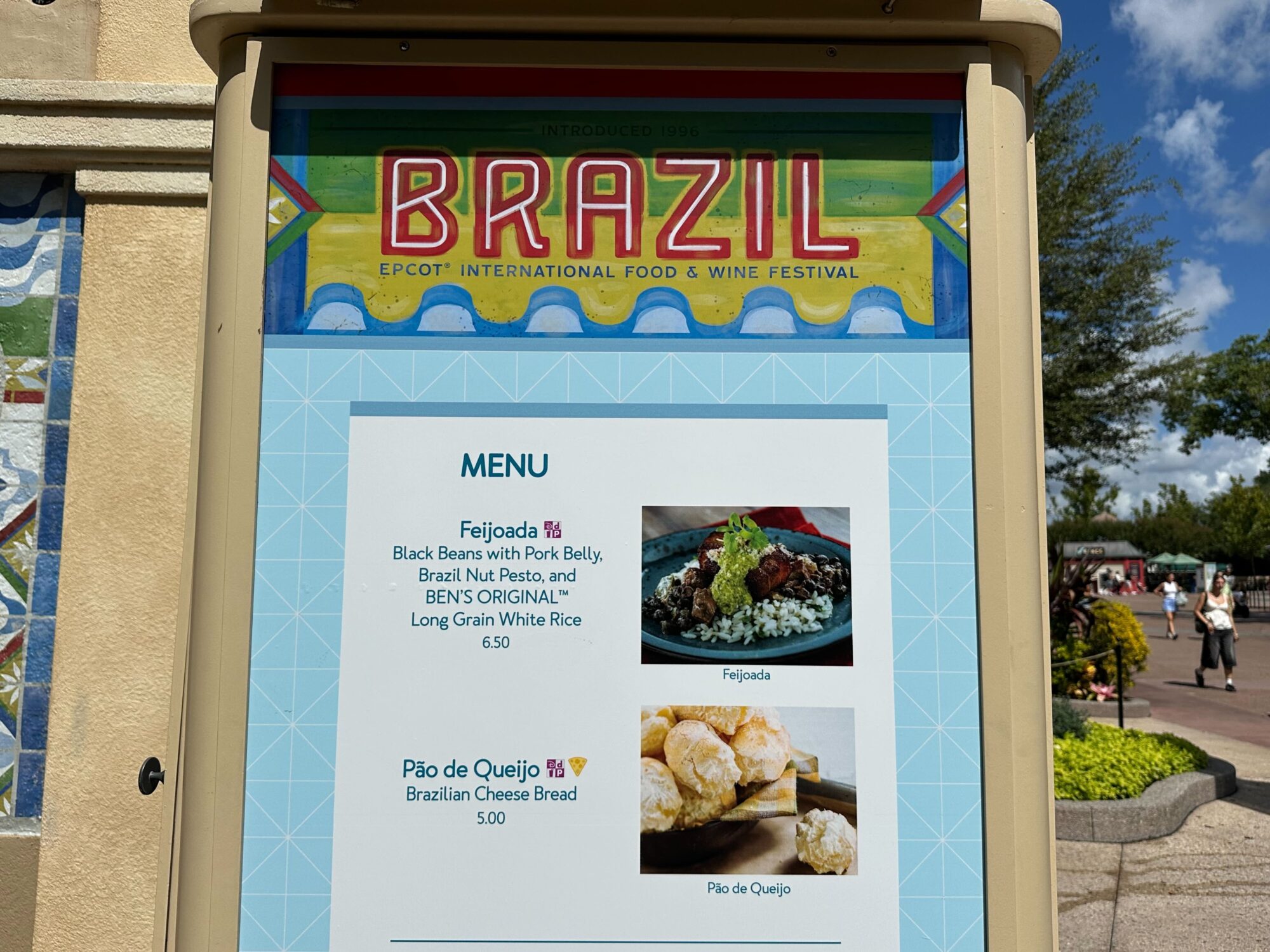Menus (With Prices) for 2024 EPCOT International Food & Wine Festival 51 A menu sign at the Brazil booth for Epcot International Food & Wine Festival displays Feijoada and Pão de Queijo, with prices and images of these delectable dishes. The menus highlight the rich cultural flavors that guests can experience.