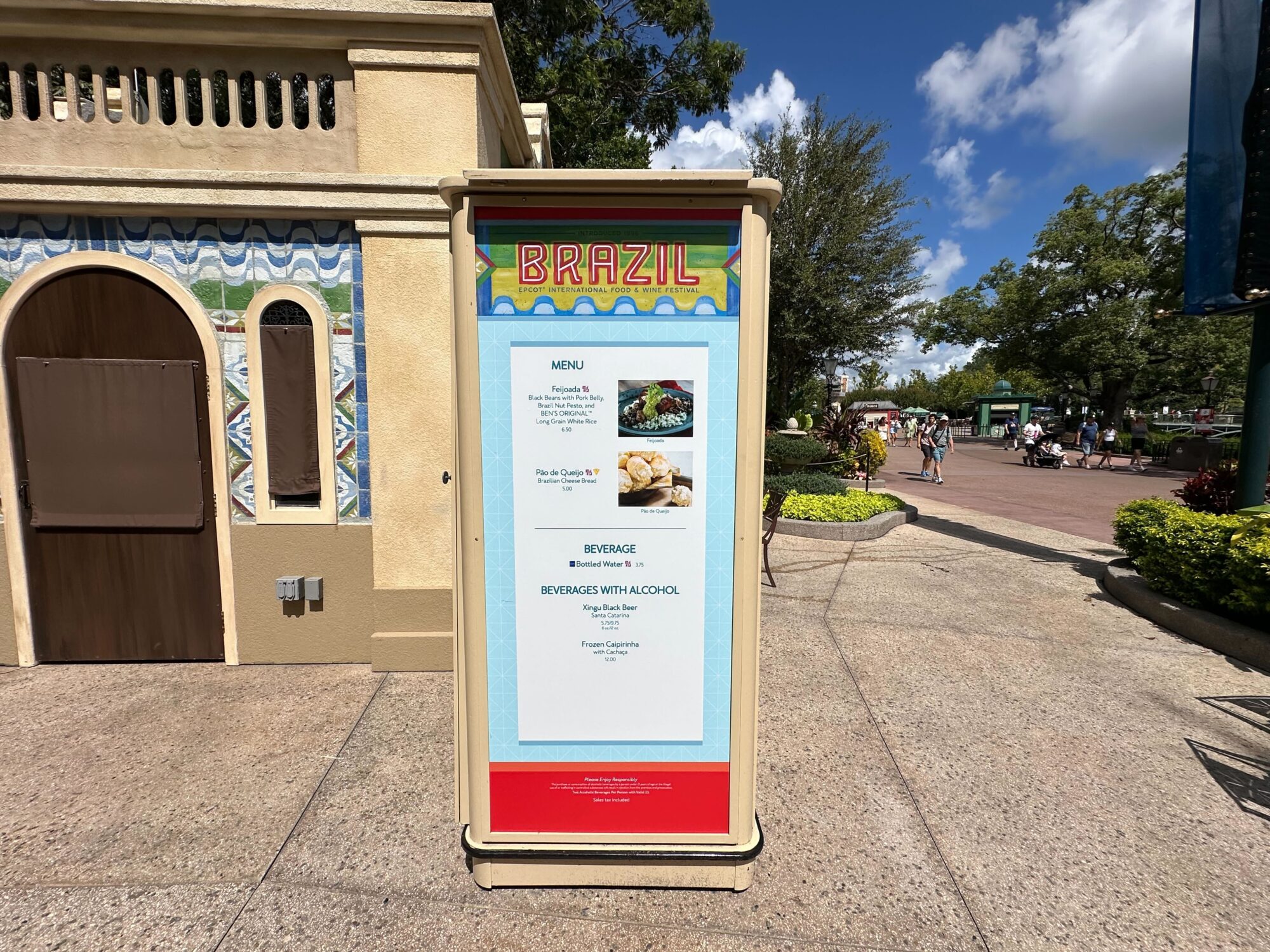 Menus (With Prices) for 2024 EPCOT International Food & Wine Festival 50 A display board at a Brazil-themed location showcases an enticing menu of items available, including food and beverages, set against lush greenery and a partly cloudy sky—perfect for a food & wine festival atmosphere.