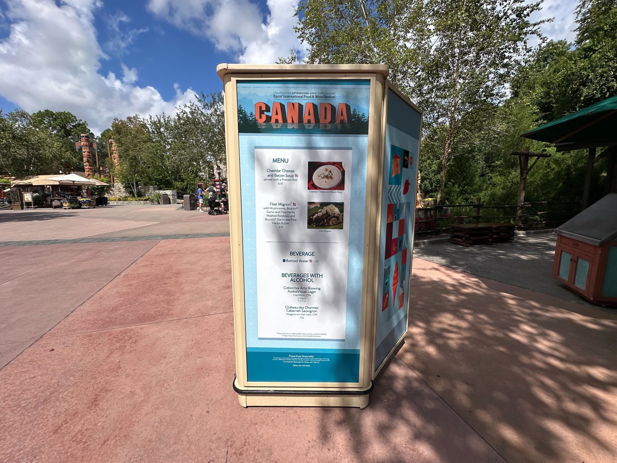Menus (With Prices) for 2024 EPCOT International Food & Wine Festival 56 A display board in a park shows the Canadian-themed menus and beverage options for a food & wine festival. The background features trees, a pathway, and other park structures, creating a scenic setting for the event.