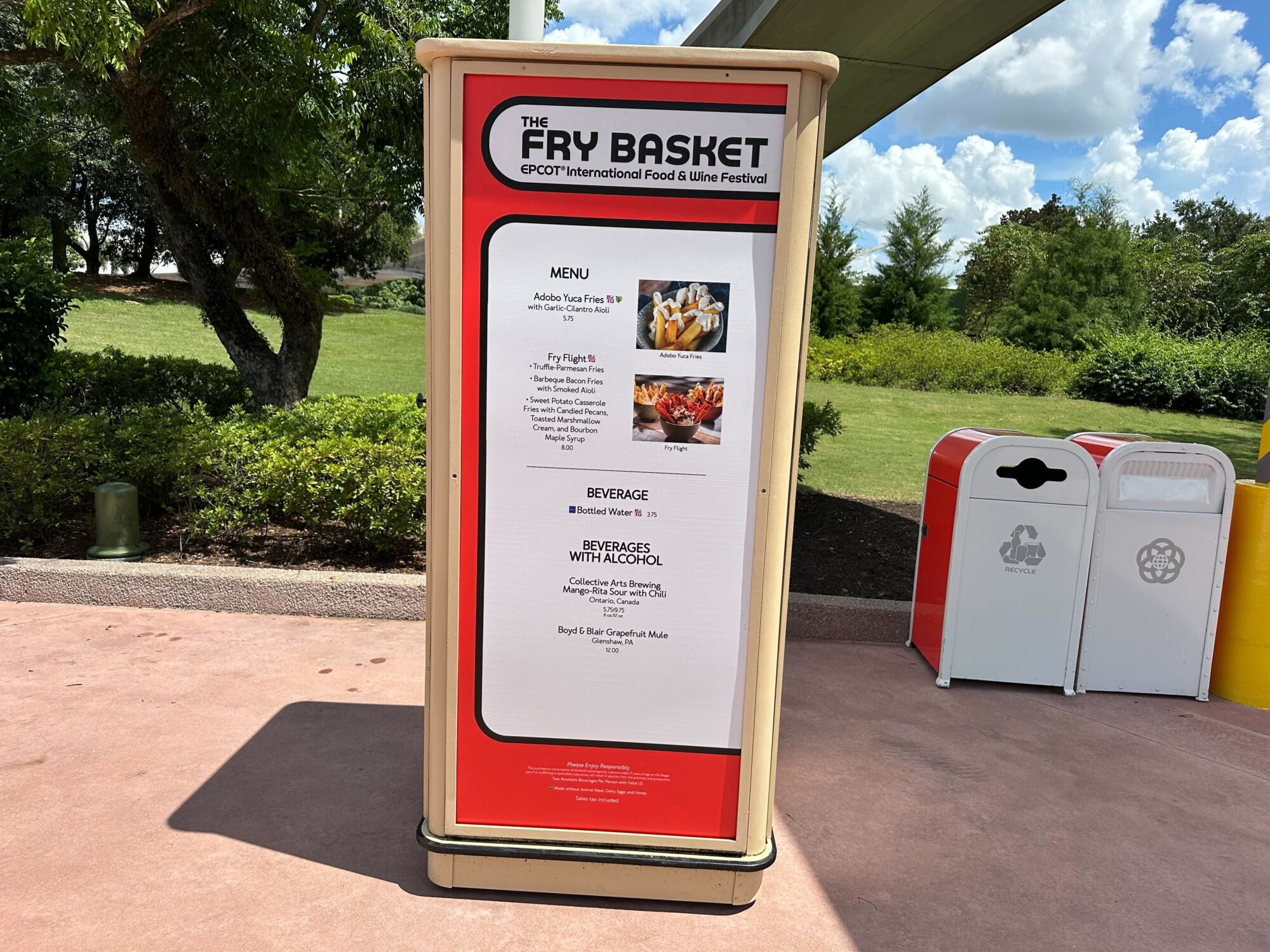 Menus (With Prices) for 2024 EPCOT International Food & Wine Festival 7 A red and white information board menus lists food and beverage options at The Fry Basket during the EPCOT International Food & Wine Festival. The board is positioned near a trash and recycling bin.