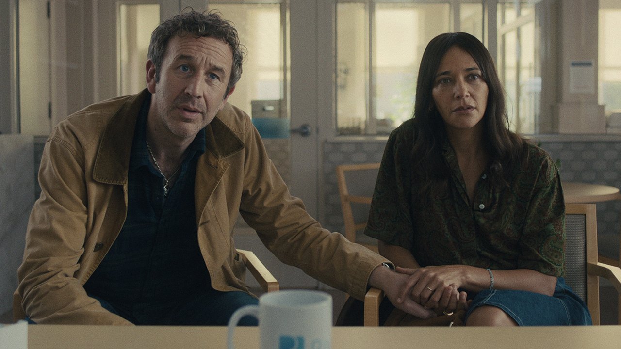 A man holds a woman's hand as they talk to someone across the table in the Common People episode of Black Mirror.