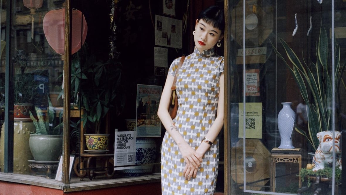 A woman wearing a patterned cheongsam standing in the doorway of a storefront.
