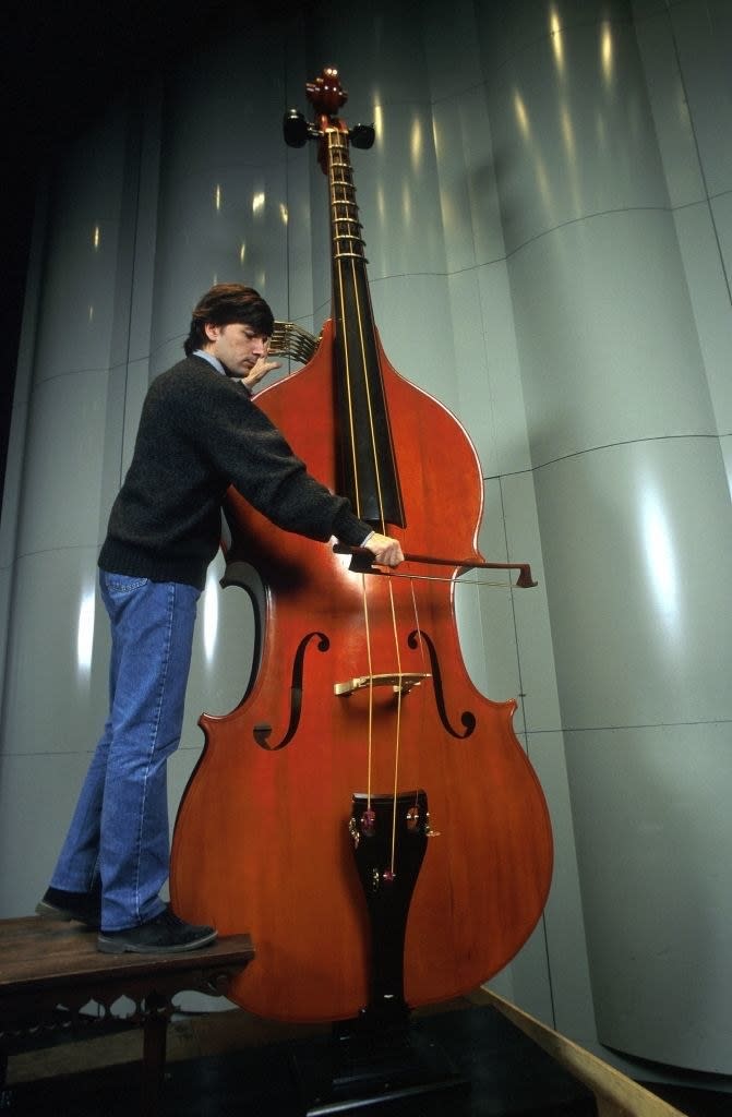 A man standing on a step alongside a huge, cello-like instrument and playing it, and it's still taller than he is