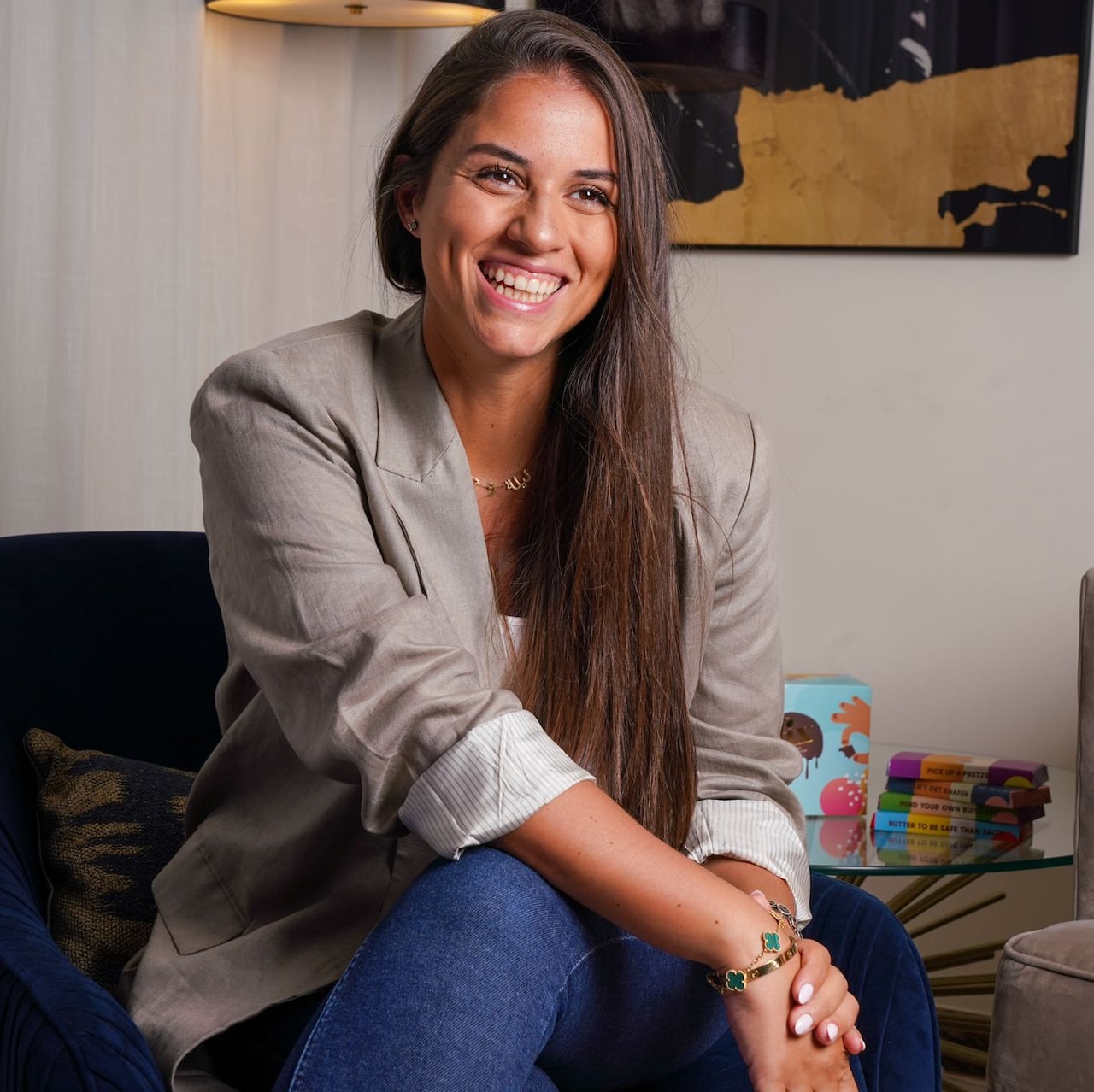 A woman in a blazer and jeans smiles while sitting in a chair