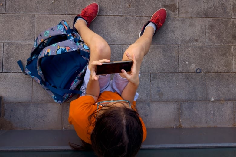 A boy with eyeglasses, wearing an orange T-shirt and red sneakers, looks at a cellphone as he sits beside his blue knapsack.