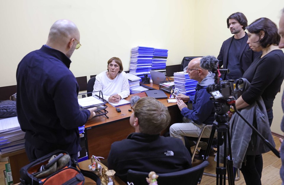 A woman sits at a desk in an office being interviewed by several people, while others look on.