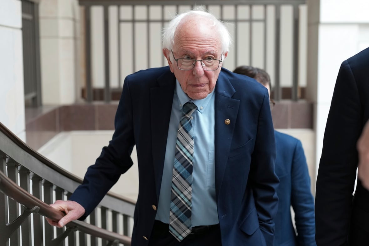 A white-haired balding, cleanshaven and bespectacled man in a suit and tie gets up to stand from a seated position.