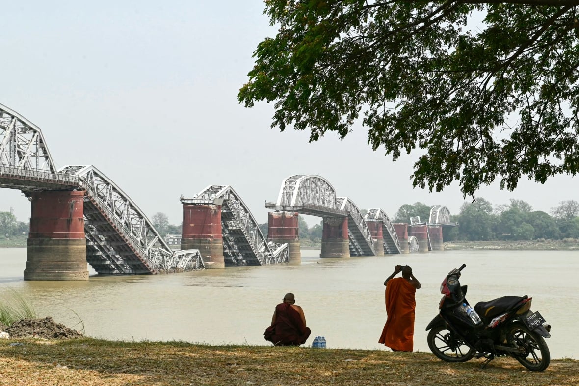 A monk in orange robes stands next to a motor scooter, on the bank of river, taking a picture of a collapsed bridge.