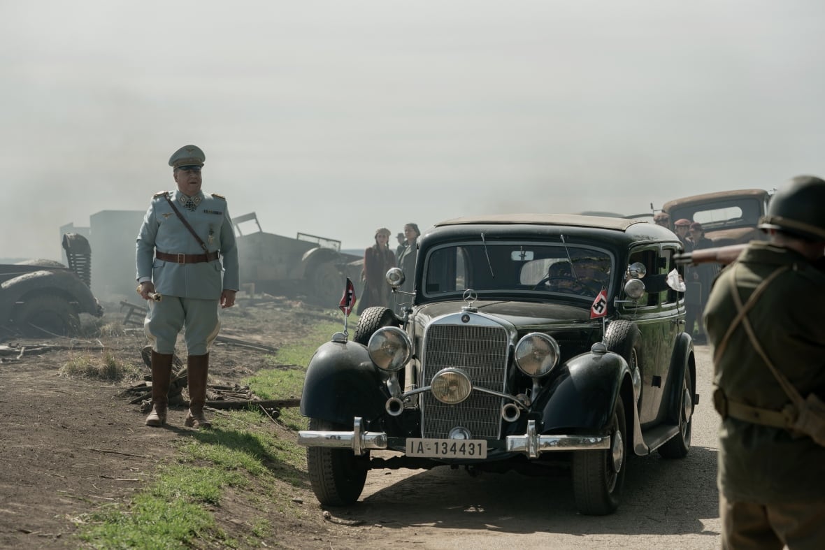A man in a military uniform walks beside an old fashioned car.