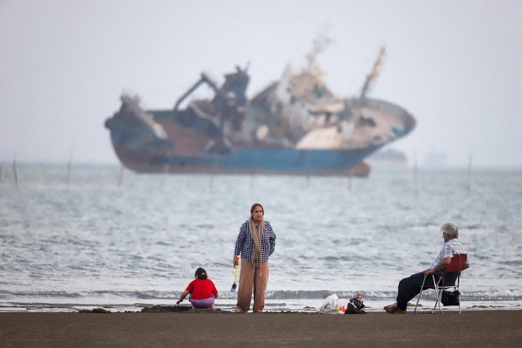 People gather on a beach, as a vessel in the Strait of Hormuz is visible, near the beach of Bandar Abbas