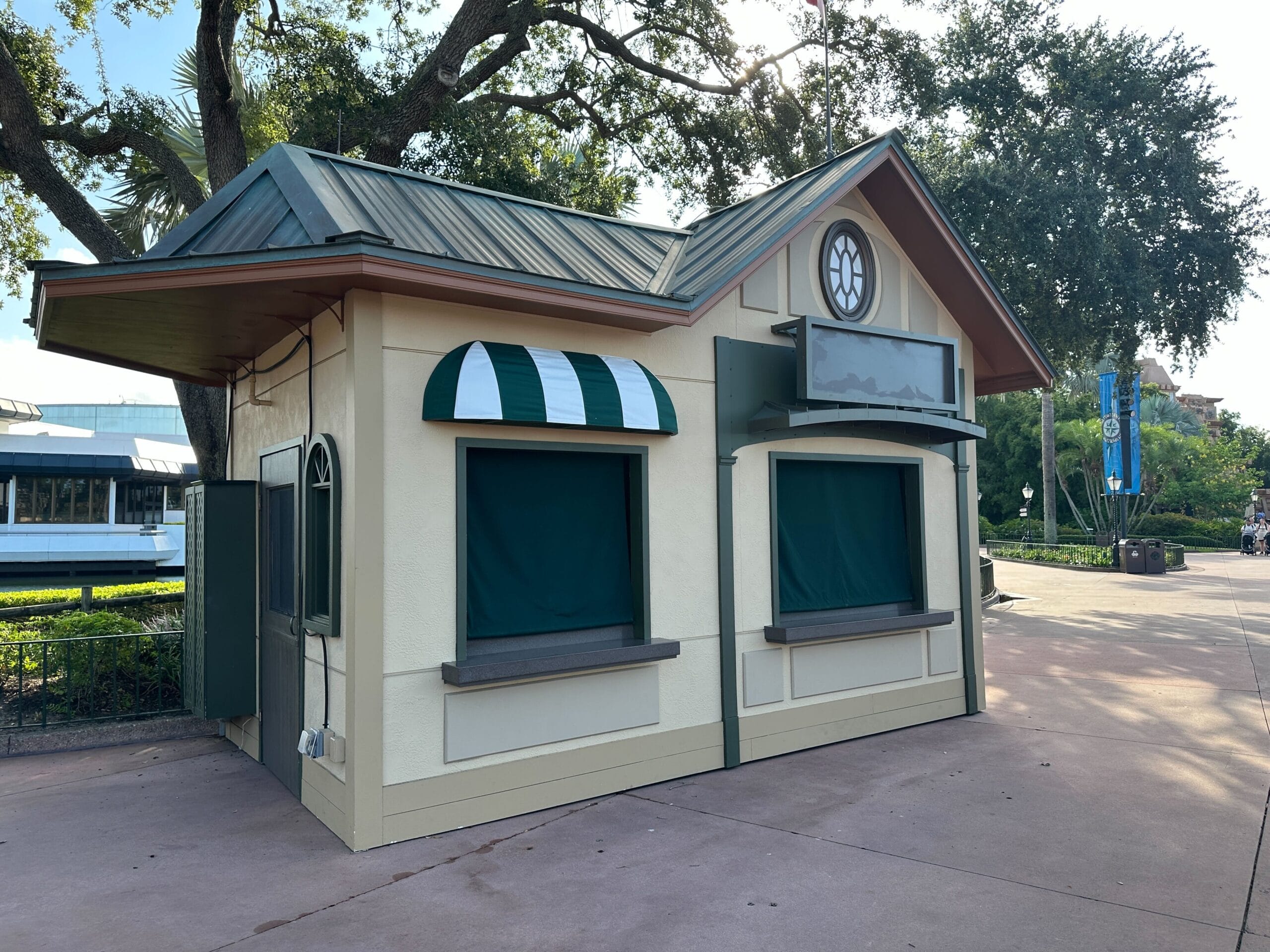 Small cream-colored kiosk with green trim and closed windows, located on a paved walkway under trees.